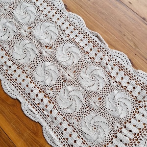 May include: A beige crocheted table runner with a delicate swirl pattern. The runner is made of cotton and has a scalloped edge.