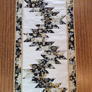 May include: A white and black table runner with a floral pattern. The runner has a gold border with a floral design. The runner is made of fabric and is approximately 14 inches wide and 36 inches long.
