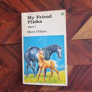 May include: A book cover featuring a blue sky background with a brown horse and a light brown foal standing in a field of green grass. The book title is "My Friend Flicka" and the author is "Mary O'Hara".