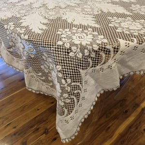 May include: An off-white lace tablecloth featuring an intricate openwork mesh pattern with detailed floral and leaf motifs. The fabric has a scalloped edge with a delicate picot trim, draping over the corner of a surface with a visible texture.