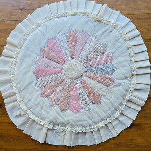 Könnte beinhalten: Ein runder, cremefarbener, gesteppter Teppich mit einem pinkfarbenen und braunen Blumendesign in der Mitte. Der Teppich hat einen weißen Spitzenrand und einen gekräuselten Rand.