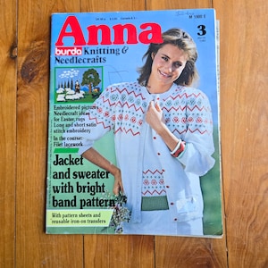 Op de afbeelding: Een vintage tijdschriftcover voor Anna Burda Breien & Handwerken, met een vrouw die een witte trui draagt met een rood-groen gepatroneerd band. De tekst op de omslag luidt "Jas en trui met een helder bandpatroon" en "Maart 1980".