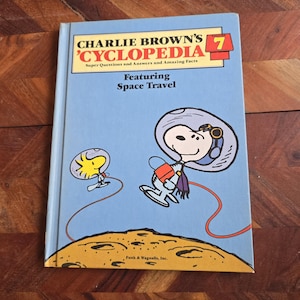 Puede incluir: Un libro vintage de tapa dura titulado "Charlie Brown's Cyclopedia Volumen 7" con Snoopy y Woodstock con trajes espaciales. La portada es azul claro con el título en amarillo y rojo, y el libro trata sobre viajes espaciales.