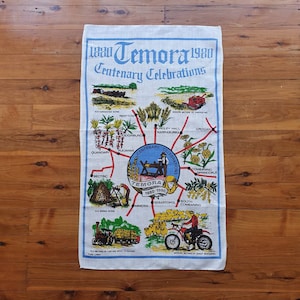 May include: A white linen tea towel with a colorful map of Temora, Australia, celebrating its centenary from 1880 to 1980. The map shows the surrounding towns and features illustrations of farming, mining, and sheep shearing.
