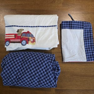 May include: A white fitted sheet with a red fire truck applique, a blue plaid sheet, and a blue plaid pillowcase. The fire truck has a dog in the driver's seat. The sheets are made of a soft, comfortable material.