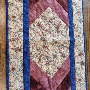May include: A quilted table runner with a floral pattern in shades of cream, brown, and blue. The runner has a diamond-shaped design with a burgundy border.