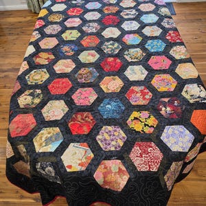 May include: A quilt featuring a repeating hexagonal pattern composed of various multicolored fabric patches with floral and abstract designs. The hexagons are separated by a black fabric border with a subtle, swirling, stitched line pattern.