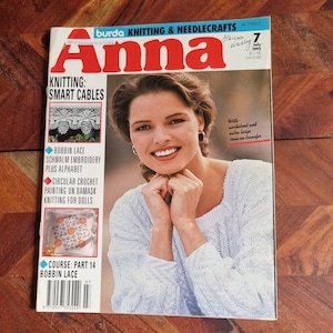以下が含まれることがあります： 1993年7月号の「アンナ」編み物と手芸雑誌の表紙。表紙には、ケーブル編みのパターンが施された白いニットセーターを着た女性が写っています。雑誌のタイトルは赤と白の文字で書かれています。表紙には、「編み物：スマートケーブル」と「ワークシートと特大アイロン転写付き」のテキストも掲載されています。