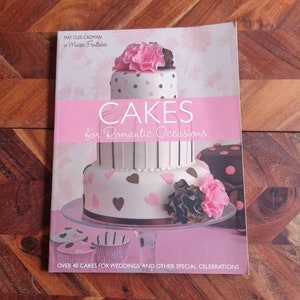 May include: A cookbook titled "Cakes for Romantic Occasions" by May Clee-Cadman of Maiste Fantaisie. The cover features a three-tiered wedding cake decorated with pink and brown frosting, hearts, and flowers.