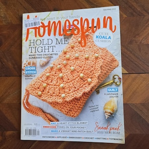 May include: Homespun magazine cover featuring a crocheted peach-coloured clutch with white beads. The magazine title is in a white font with a red outline. The cover also features a patchwork quilt and a cute koala cushion.