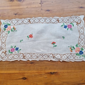 Op de afbeelding: Rechthoekige, gebroken witte geborduurde tafelloper met een geschulpte rand. De loper heeft kleurrijke bloemenborduurwerk in roze, blauw, oranje en rood. Een bruin geometrisch patroon omlijst de randen.
