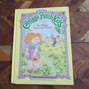 May include: A yellow book cover featuring a green and purple illustration of a Cabbage Patch Kid. The title of the book is "Cabbage Patch Kids" and the subtitle is "The Shyest 'Kid in the 'Patch". The book is a Parker Brothers Story Book, written by Mark Taylor and illustrated by Lynn Lace.