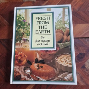 Op de afbeelding: Een wit kookboek met de titel "Fresh From The Earth: The Four Seasons Cookbook". De omslag toont een verscheidenheid aan voedingsmiddelen, waaronder een glas sinaasappelsap, een kom met fruit, een dessert met een romige topping en een gebakje.