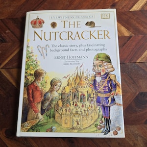 Könnte beinhalten: Ein gebundenes Buch mit dem Titel "The Nutcracker" von Ernst Hoffmann, illustriert von James Mayhew. Das Cover zeigt Illustrationen von Figuren und Szenen aus der Geschichte, darunter ein Nussknacker und ein Weihnachtsbaum.