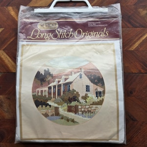 Puede incluir: Un kit SEMCO Long Stitch Originals con una escena de cabaña. El diseño, en un marco circular, representa una casa con porche, rodeada de árboles y una valla. El kit está empaquetado en una bolsa de plástico transparente con asa blanca.
