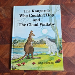 May include: A book cover with a blue sky and green grass background. The title is "The Kangaroo Who Couldn't Hop and The Cloud Wallaby" by Pixie O'Harris. A brown kangaroo and a white wallaby are standing on the grass.