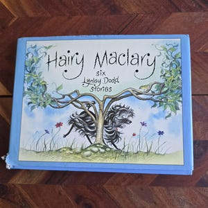 May include: A blue book cover with the title "Hairy Maclary" in cursive script. The book cover features an illustration of two black dogs running through a tree with green leaves and blue flowers.