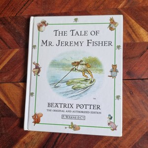 May include: A white book cover with a green border. The title is "The Tale of Mr. Jeremy Fisher" by Beatrix Potter. The cover features an illustration of a frog in a boat on a lake.