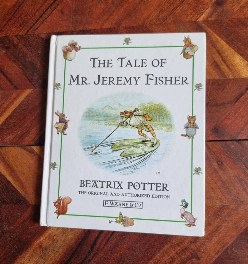 May include: A white book cover with a green border. The title is "The Tale of Mr. Jeremy Fisher" by Beatrix Potter. The cover features an illustration of a frog in a boat on a lake.