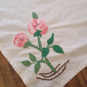 以下が含まれることがあります： 白い布地に、ピンクの花と緑の葉が刺繍されています。花はピンク色の芯を持ち、緑の葉で囲まれています。葉は緑色の糸で縫われています。花は茶色の茎でつながっています。茎は茶色の糸で縫われています。布地は白で、滑らかな質感です。