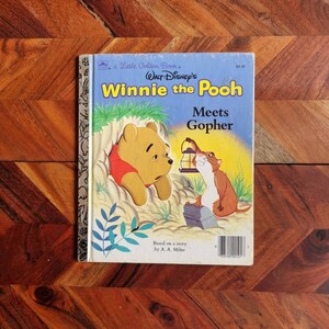 Könnte beinhalten: Ein Vintage-Buch aus der Little Golden Book-Reihe mit Winnie Puuh, einem beliebten Comic-Bären in einem gelben und roten Outfit. Der Buchumschlag zeigt Winnie Puuh, der aus einem Loch im Boden schaut, während ein Gopher eine Laterne hält. Der Titel lautet "Winnie Puuh trifft Gopher" und das Buch basiert auf einer Geschichte von A. A. Milne.