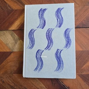 Puede incluir: Una cubierta de libro azul claro con un patrón repetitivo de líneas onduladas azules. Las líneas están pintadas a mano y tienen un aspecto texturizado.