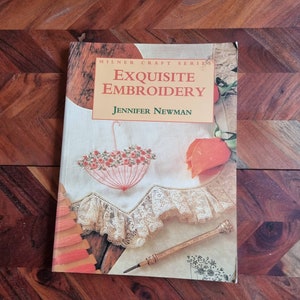 May include: A book titled "Exquisite Embroidery" by Jennifer Newman. The book cover features a white fabric with an embroidered umbrella and a rose. The book is part of the Milner Craft Series.