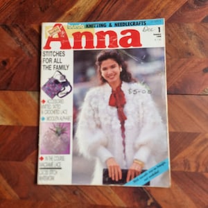 Op de afbeelding: Een tijdschriftcover voor Anna, een Burda brei- en handwerkmagazine. De cover toont een vrouw die een witte gebreide cardigan draagt met een rode strik. De titel van het tijdschrift is in rood-witte letters. De tekst op de cover luidt "Steken voor het hele gezin".