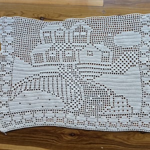 May include: A rectangular off-white crochet doily featuring a detailed filet pattern that depicts a large sailing ship on the water with a sun in the upper right corner. The intricate needlework creates a grid-like texture throughout the piece.