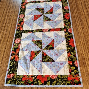 May include: A blue and white quilted table runner with a geometric pattern and a black, green and red border. The border features a festive design with holly, poinsettias and bows.