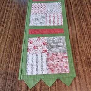 May include: A green table runner with a scalloped edge. The runner is made of fabric and has a patchwork design with different floral patterns in shades of red, white, and grey.