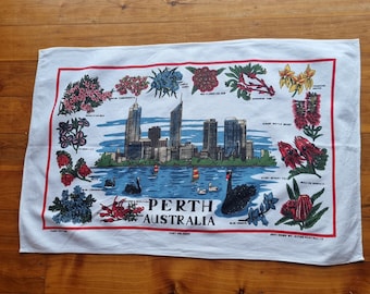 Op de afbeelding: Een witte theedoek met een kleurrijke illustratie van de skyline van Perth en inheemse Australische bloemen. De tekst "PERTH AUSTRALIA" is in zwarte letters onder de skyline gedrukt.