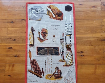 Op de afbeelding: Een rode en witte theedoek met een ontwerp van Aboriginal kunst uit Australië. Het ontwerp omvat een gedecoreerde boemerang, een gesneden figuur, een vis, een speer, een boodschapstok en een handtekening. De tekst op de theedoek luidt "Greetings from Lake Keepit N.S.W. Aboriginal Art Australia".