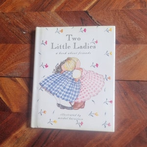 May include: A white book with a colorful illustration of two girls hugging. The book title is "Two Little Ladies" and the subtitle is "a book about friends". The book is illustrated by Michel Bernstein.