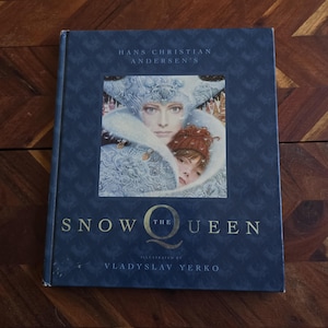 May include: A hardcover book titled Hans Christian Andersen's The Snow Queen, illustrated by Vladyslav Yerko. The dark blue cover features a central illustration of a pale figure in an ornate white headpiece embracing a child with reddish hair.