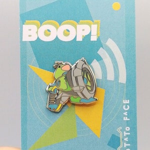 May include: A green and gray enamel pin featuring a cartoon character riding a motorcycle. The pin says "BOOP!" and "POTATO FACE" on the background.