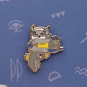 Puede incluir: Un pin metálico con un búho sosteniendo un arma. El búho es blanco y gris, con detalles amarillos en el arma. El pin está sobre un fondo azul con varios símbolos.