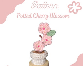 May include: A crocheted potted cherry blossom plant. The plant features pink flowers with yellow centers, green leaves, and a brown stem, all in a white pot on a wooden base. The text "Pattern Potted Cherry Blossom" is displayed in pink.
