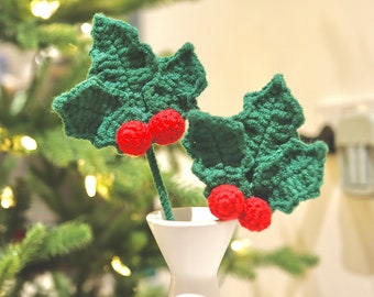 May include: Two crocheted holly sprigs with red berries in a white vase on a white table. The sprigs are green and the berries are red.