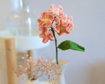 May include: A pink crocheted flower bouquet in a white vase on a white table. The bouquet is made of several pink flowers with yellow centers. The flowers are arranged in a cluster and have a green leaf.