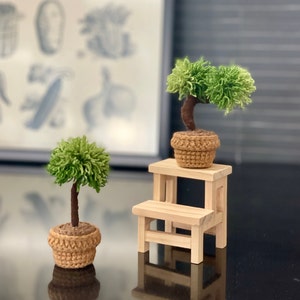 May include: Two small potted trees with green yarn foliage and brown yarn pots. The trees are on a wooden step stool with a light wood finish.