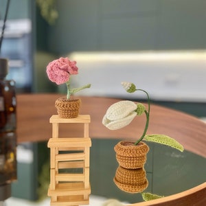 Può includere: Due composizioni floreali all'uncinetto in vasi marroni. Un fiore è rosa e l'altro è bianco. Il fiore rosa è su un piccolo scaffale di legno.