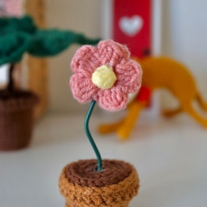 May include: A small crocheted pink flower with a yellow center is attached to a green wire stem and placed in a brown crocheted pot.