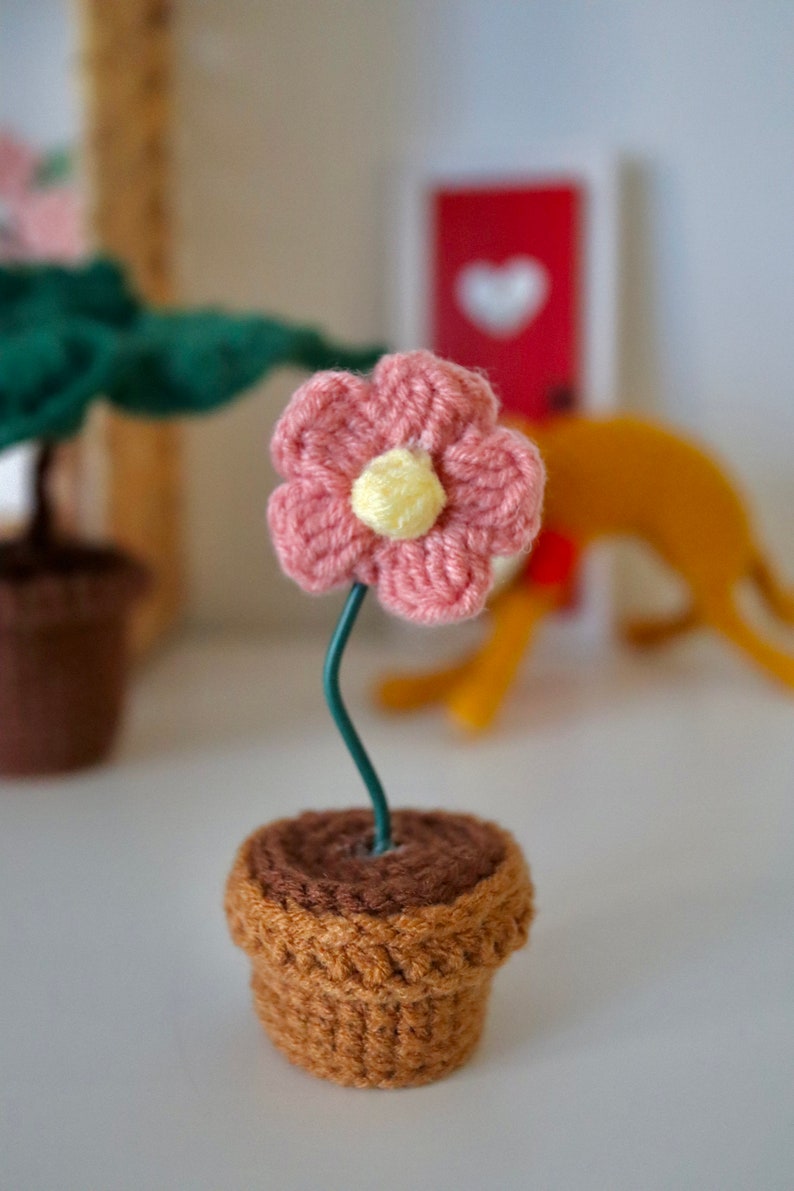 May include: A small crocheted pink flower with a yellow center is attached to a green wire stem and placed in a brown crocheted pot.