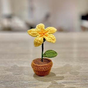 May include: A crocheted yellow flower with an orange centre, a green leaf, and a brown pot. The flower is sitting on a wooden surface.