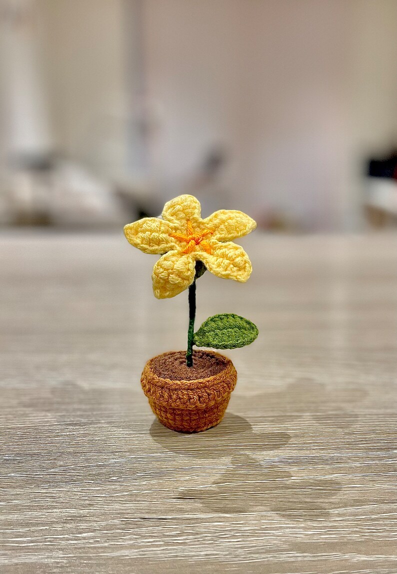 May include: A crocheted yellow flower with an orange centre, a green leaf, and a brown pot. The flower is sitting on a wooden surface.