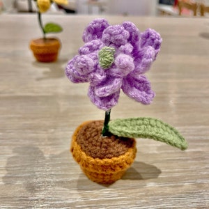 May include: A crocheted purple flower with a green leaf in a brown and orange pot.
