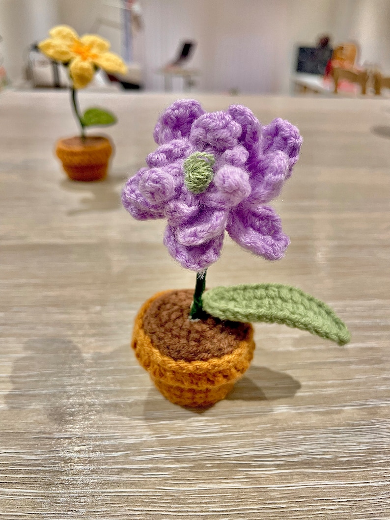 May include: A crocheted purple flower with a green leaf in a brown and orange pot.