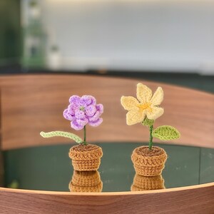May include: Two crocheted flower pots, one with a purple flower and the other with a yellow flower. The pots are brown and the flowers are sitting on a wooden tray.