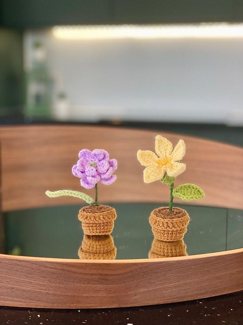 May include: Two crocheted flower pots, one with a purple flower and the other with a yellow flower. The pots are brown and the flowers are sitting on a wooden tray.